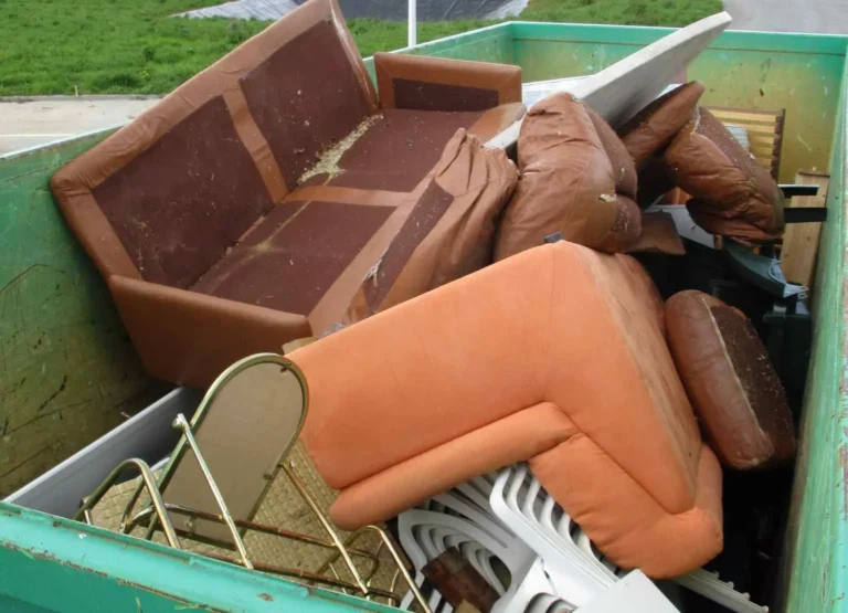 Une benne remplie de meubles mis au rebut, y compris des canapés, des coussins, des chaises et divers articles ménagers.