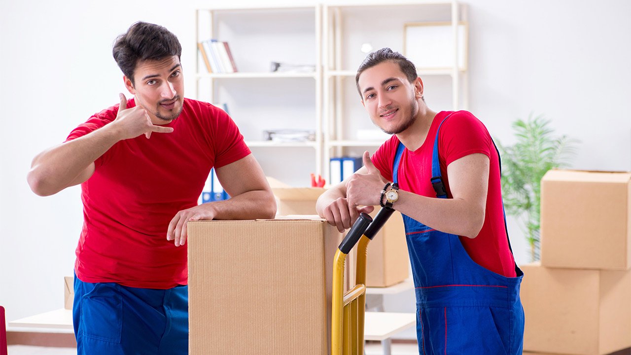 Deux hommes avec une boîte en carton dans un bureau ; l'un fait un geste pour téléphoner, l'autre lève le pouce, avec des boîtes en mouvement à l'arrière-plan.