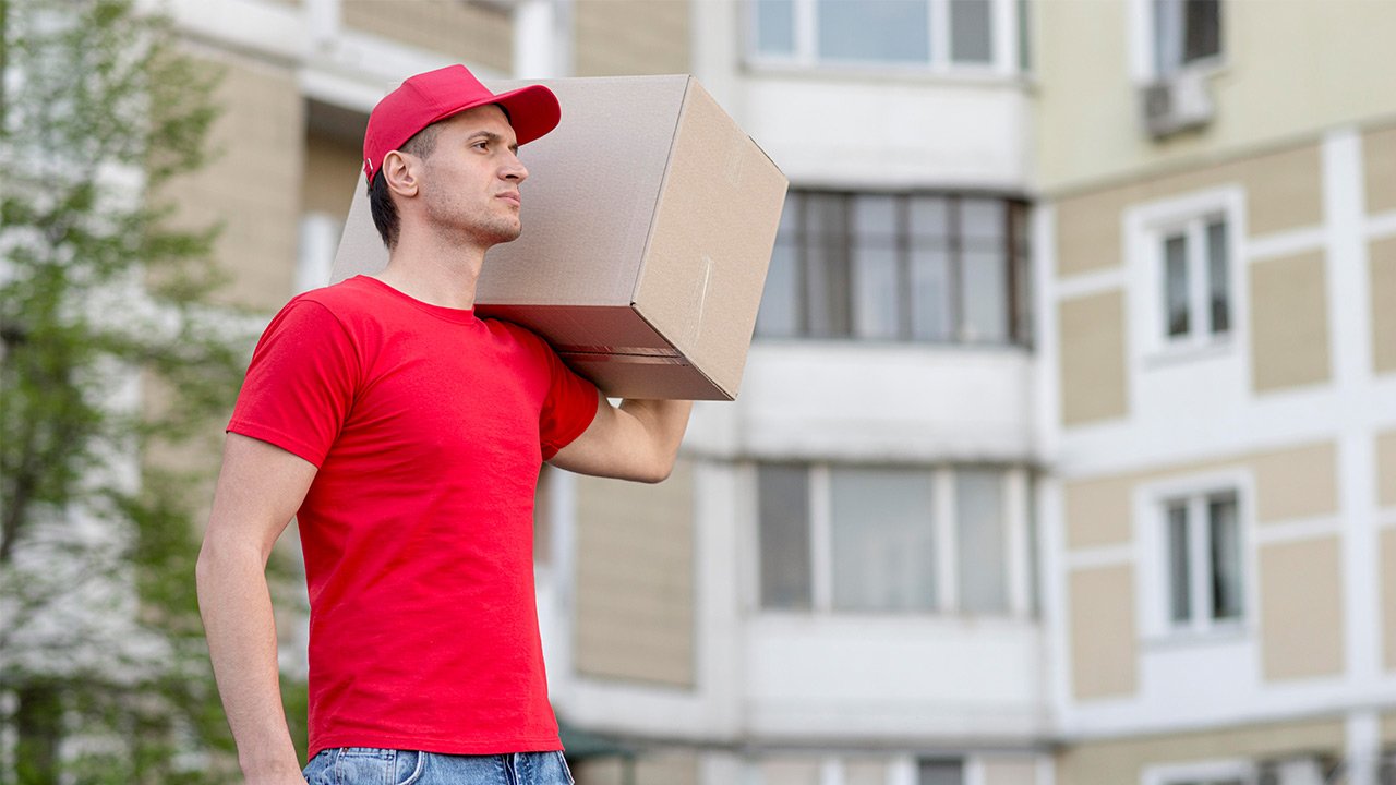 Un livreur en chemise rouge et casquette porte un carton sur l'épaule à l'extérieur d'un immeuble d'habitation.