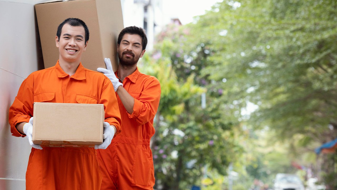 Deux hommes en uniforme orange et gants blancs transportent des cartons à l'extérieur par une journée ensoleillée, avec de la verdure et un trottoir en arrière-plan.