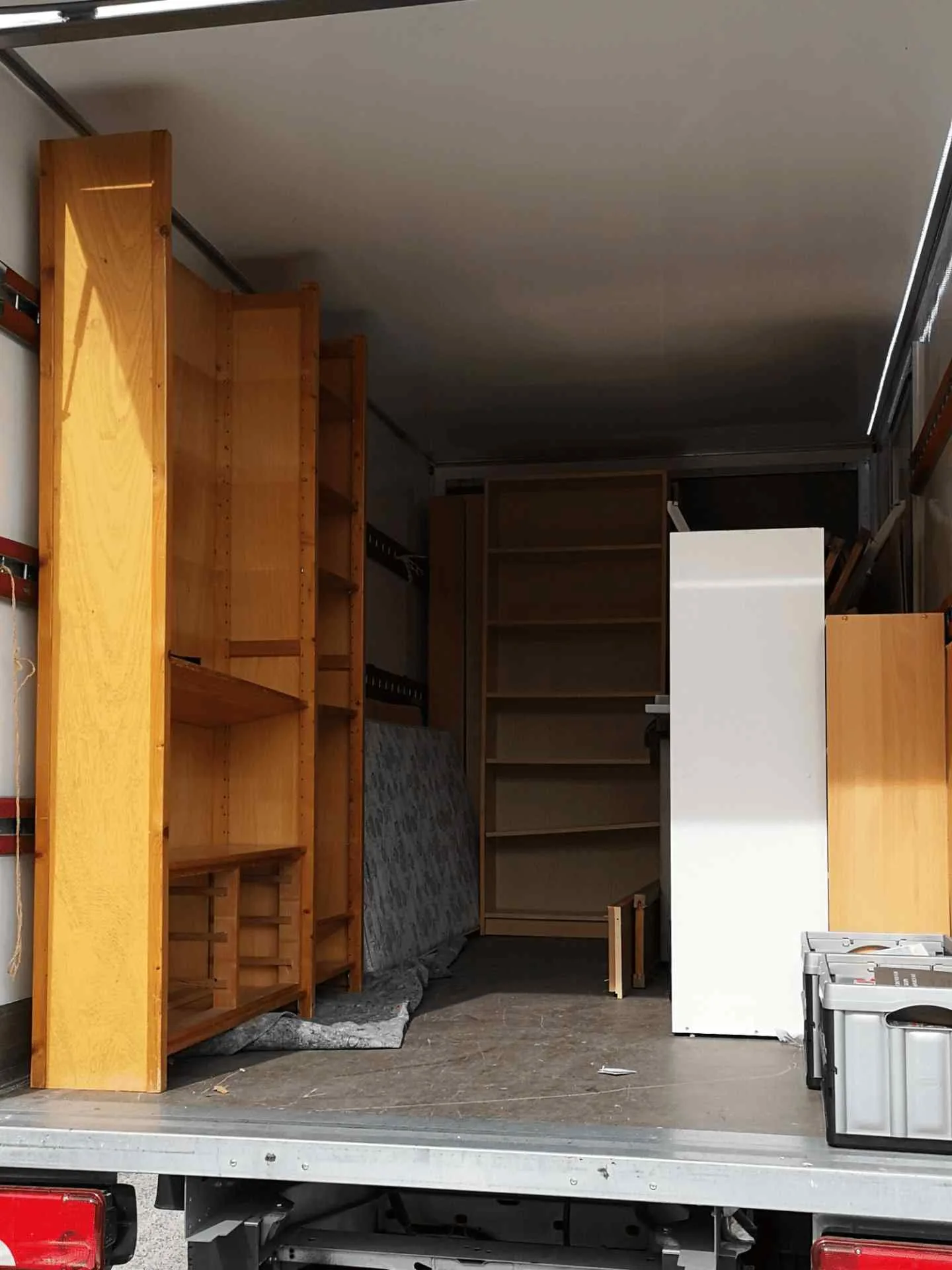 Plusieurs étagères et meubles en bois sont chargés debout à l'arrière d'un camion de déménagement, avec des cartons et d'autres objets placés autour d'eux.