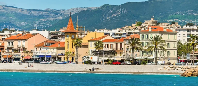 Charmante ville côtière de Cagnes sur Mer avec une plage, des bâtiments colorés, une tour d'horloge, des palmiers et des montagnes en arrière-plan sous un ciel bleu.