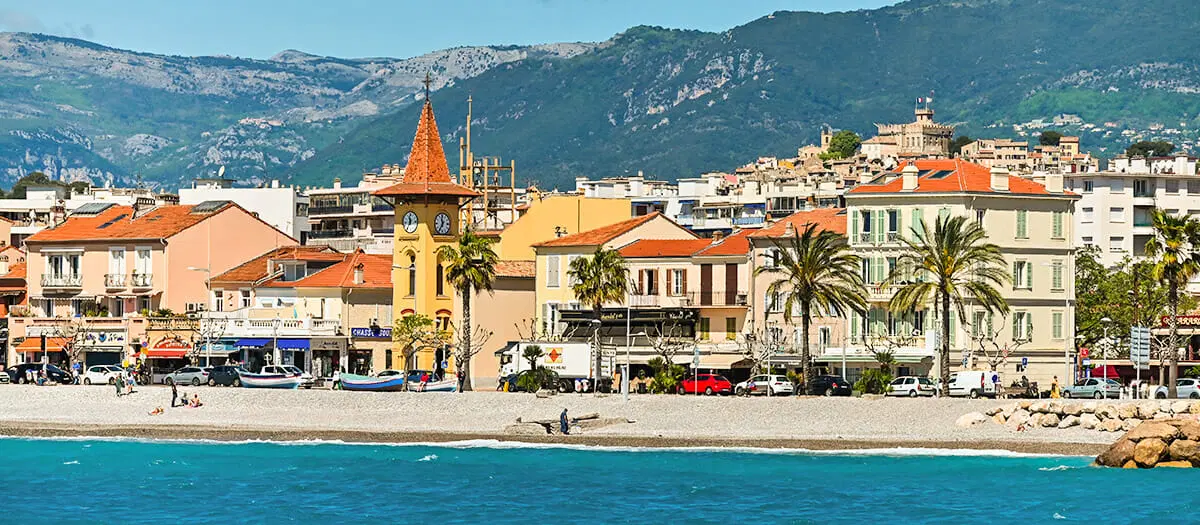Charmante ville côtière de Cagnes sur Mer avec une plage, des bâtiments colorés, une tour d'horloge, des palmiers et des montagnes en arrière-plan sous un ciel bleu.