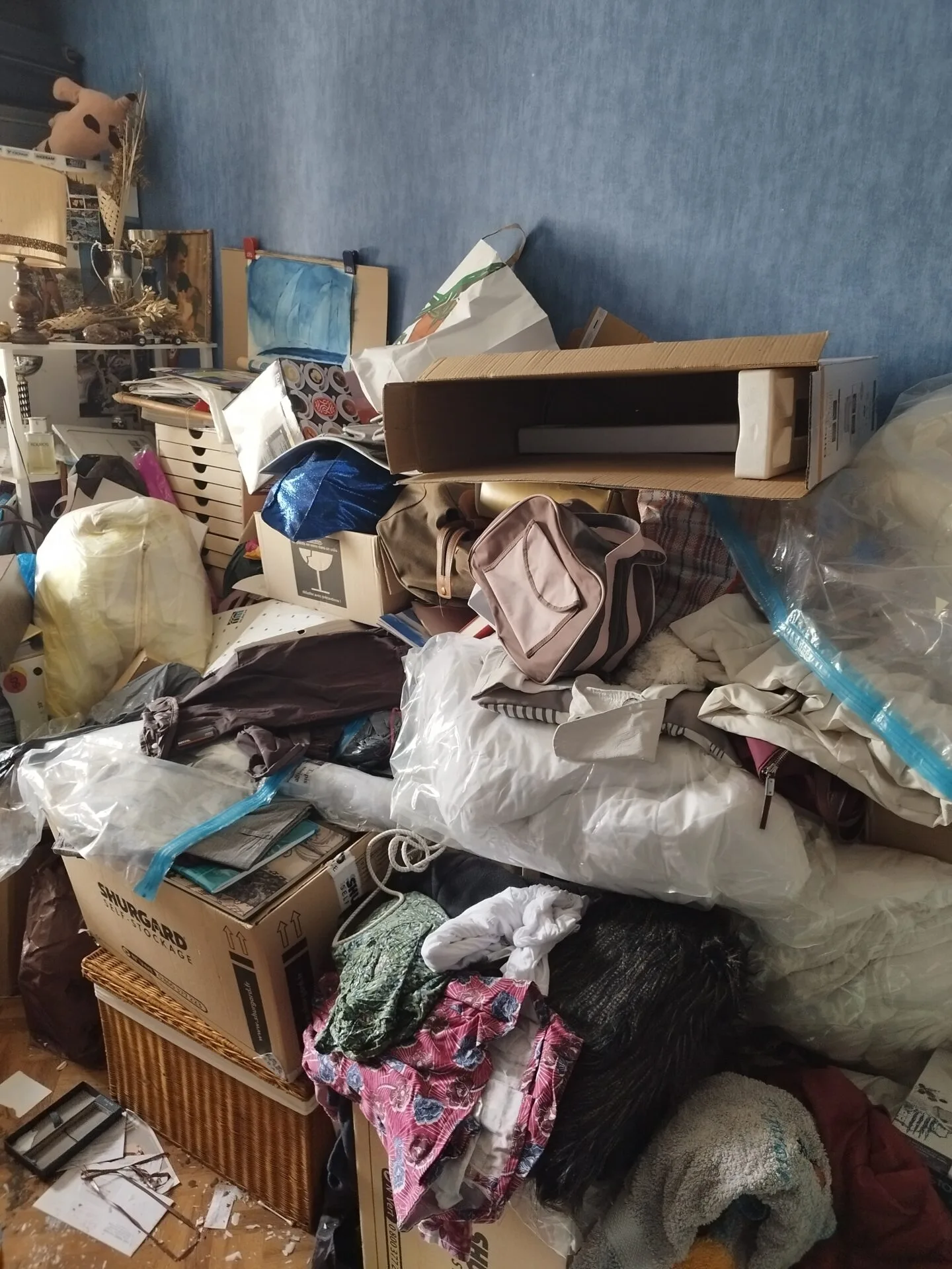 Une pièce encombrée de vêtements, de boîtes, de sacs et d'objets ménagers divers empilés les uns sur les autres contre un mur bleu suggère un besoin de débarras ou peut même indiquer un syndrome de Diogène.