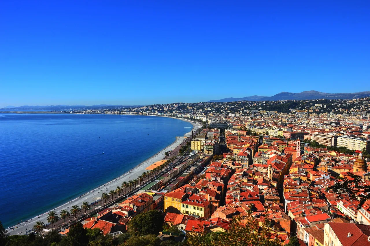 Vue aérienne d'une ville côtière avec des bâtiments aux toits rouges, une plage incurvée, des palmiers et des collines en arrière-plan sous un ciel bleu clair.
