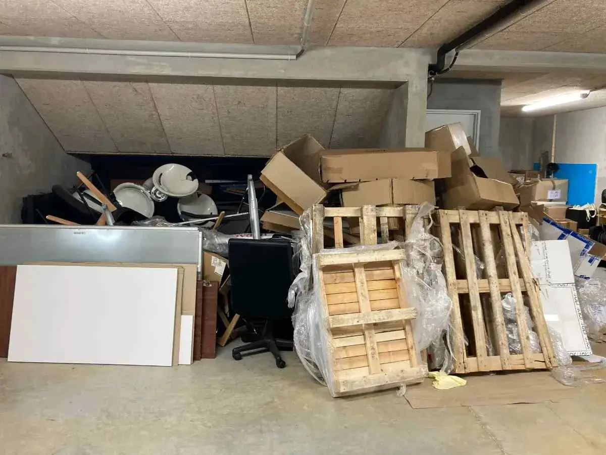 Un espace de stockage encombré de palettes en bois, de cartons, de tambours, d'une chaise de bureau et de divers matériaux empilés de manière désordonnée contre un mur.