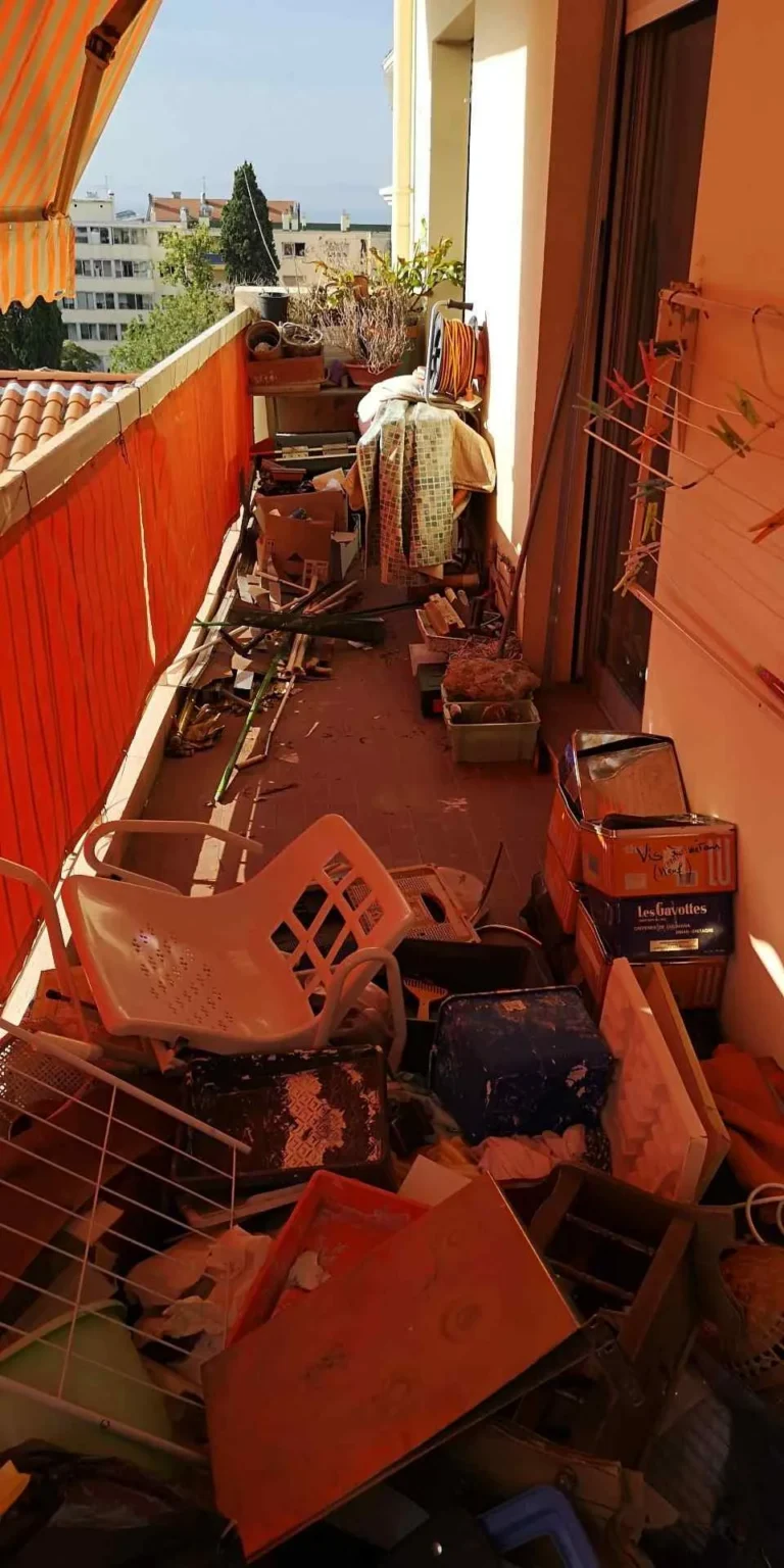 Un balcon encombré de divers objets, dont des cartons, une chaise cassée, un séchoir et des débris épars, partiellement ombragé par un auvent orange.
